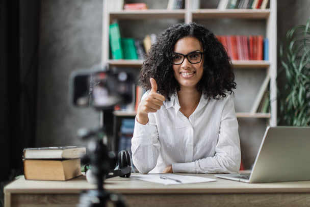 Smiling multiracial woman in white shirt sitting at desk with books, notes and laptop recording video blog and showing thumbs up. Female tutor working at home during distance learning. - Foto, immagini