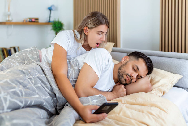 Žena žárlí a je podezřívavá a špehuje svého parťáka v jeho smartphonu, když spí v ložnici. Manželka špehuje manželův telefon, zatímco spí. Koncept nedůvěry, žárlivosti - Fotografie, Obrázek