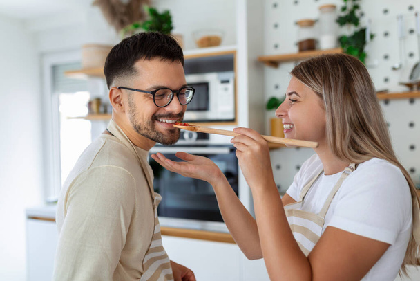 Beautiful young couple is looking at each other and feeding each other with smiles while cooking in kitchen at home. Loving joyful young couple embracing and cooking together, having fun in the kitchen - Photo, Image