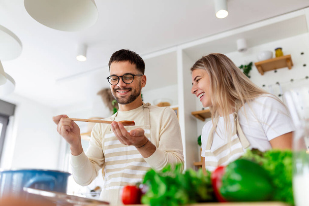 Evdeki mutfakta birlikte yemek pişiren mutlu genç çiftin portresi. Romantik genç kadın ve yakışıklı erkek aydınlık modern mutfakta dururken birlikte vakit geçirmekten zevk alıyorlar.. - Fotoğraf, Görsel