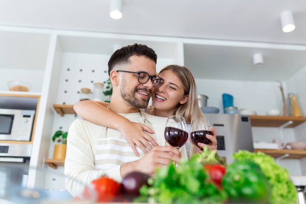 Lächelndes junges Paar, das gemeinsam in der Küche Essen kocht und eine tolle Zeit miteinander verbringt. Mann und Frau lachen und trinken Wein - Foto, Bild