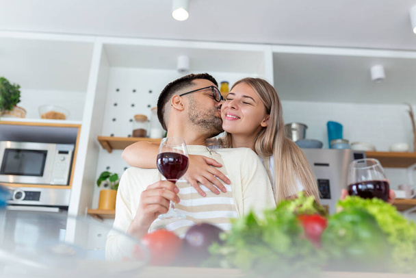 Szeretetteljes fiatalember megcsókolja a feleségét, miközben együtt főznek. Gyönyörű fiatal pár beszél és mosolyog, miközben főzés egészséges étel a konyhában otthon.Man csókolja a barátnője arcán - Fotó, kép