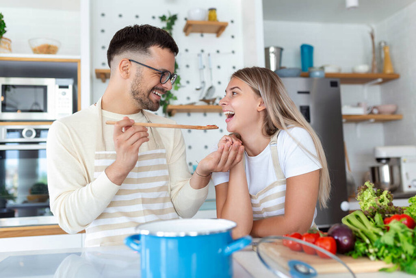 Krásný mladý pár se na sebe dívá a krmí se navzájem úsměvy, zatímco vaří doma v kuchyni. Milující radostný mladý pár objímající a vařící společně, baví v kuchyni - Fotografie, Obrázek