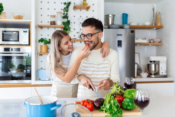 Gyönyörű fiatal pár eteti egymást, és mosolyog főzés közben otthon a konyhában. A boldog pár egészséges ételeket készít a világos konyhán. Egészséges élelmiszer-koncepció. - Fotó, kép