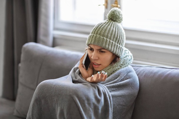 Παγωμένη νεαρή γυναίκα τυλιγμένη σε κουβέρτα κάνοντας τηλεφώνημα στο σπίτι, στρεσαρισμένη γυναίκα ενοικιαστής κάθεται στον καναπέ και μιλώντας στο κινητό, διαμαρτύρονται για την κρύα θερμοκρασία στο διαμέρισμα, closeup - Φωτογραφία, εικόνα
