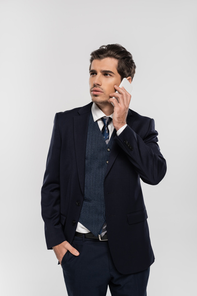 hombre de negocios confiado en traje de pie con la mano en el bolsillo y hablando en el teléfono inteligente aislado en gris  - Foto, Imagen