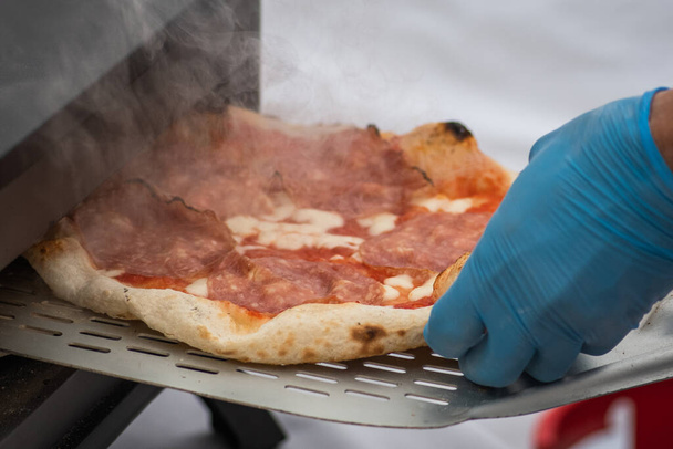 Plastik eldiven ile pişirme veya pişirme domates soslu sıcak pizza, mozzarella ve elektrikli ya da gaz fırınında kürekle salam eti, dumanla yakın çekim - Fotoğraf, Görsel