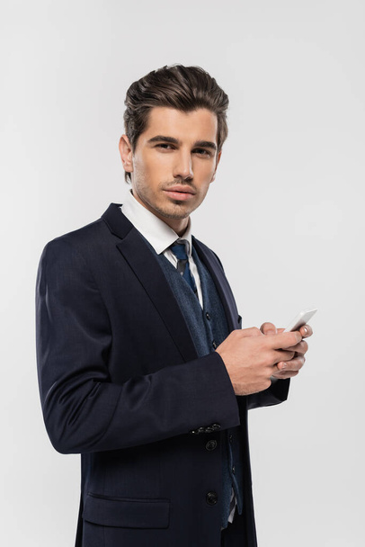 young and successful businessman in suit messaging on smartphone isolated on grey - Photo, Image