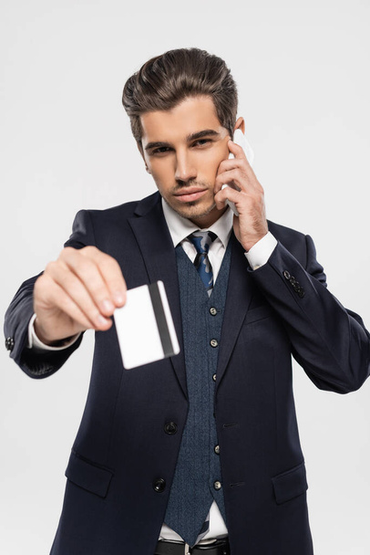 hombre de negocios seguro de sí mismo hablando por teléfono inteligente y manteniendo la tarjeta de crédito aislada en gris  - Foto, imagen