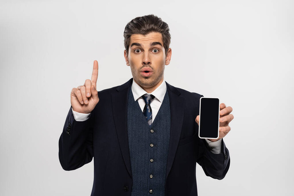 surprised businessman in suit holding smartphone with blank screen and pointing up isolated on grey  - Foto, Imagem