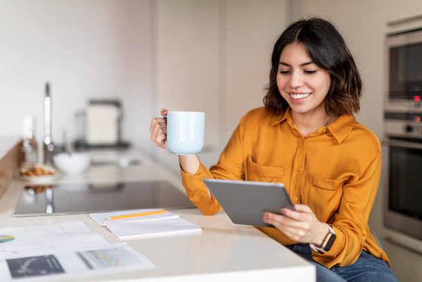 Smiling arab woman drinking coffee and using digital tablet in kitchen at home, young middle eastern female freelancer surfing internet on tablet computer and enjoying hot drink, copy space - Photo, Image