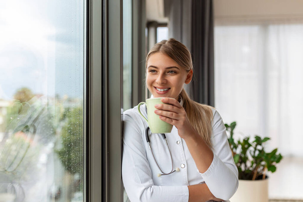 Aufnahme einer Ärztin, die eine Tasse Kaffee in der Hand hält, während sie in der Sprechstunde nach vorne blickt. Lächelnde junge Ärztin in weißer Arztuniform und Stethoskopblick in der Fernplanung. - Foto, Bild