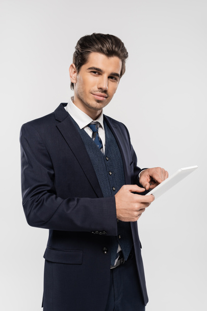 alegre joven hombre de negocios en traje sosteniendo tableta digital aislado en gris  - Foto, Imagen
