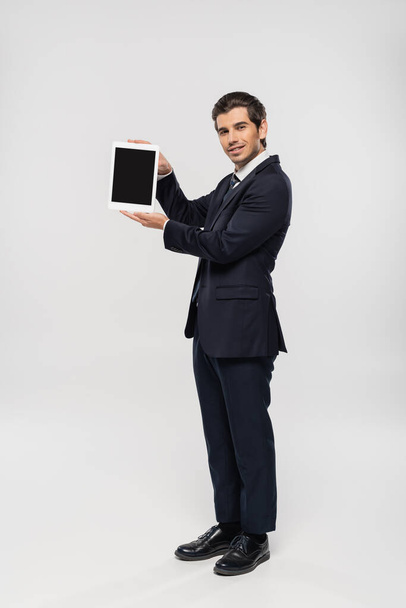 full length of cheerful businessman in suit holding digital tablet with blank screen on grey  - Φωτογραφία, εικόνα