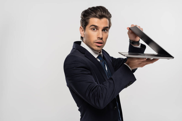 stunned businessman in suit looking at camera and holding laptop isolated on grey - Photo, Image