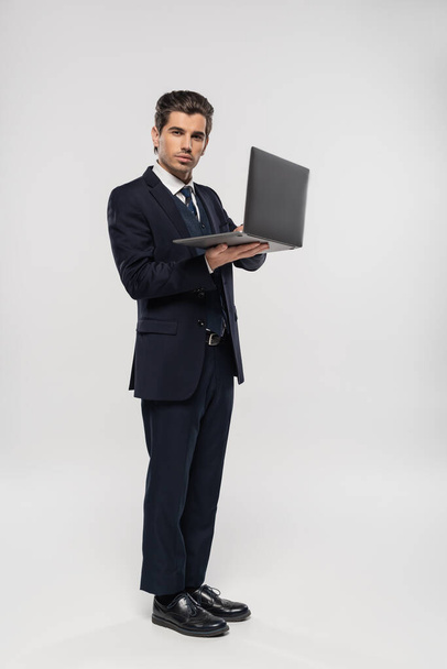 full length of confident businessman in suit using laptop while working remotely on grey - Photo, Image