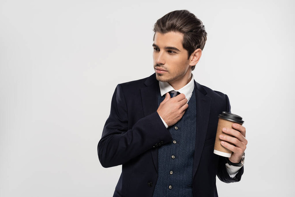 young businessman holding paper cup while adjusting tie isolated on grey - Fotografie, Obrázek