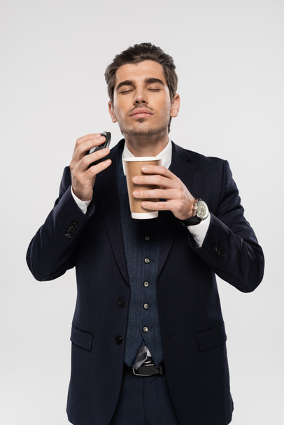 επιχειρηματίας με κλειστά μάτια κρατώντας χάρτινο κύπελλο και καπάκι, ενώ μυρίζει πρωινό καφέ απομονωμένο σε γκρι - Φωτογραφία, εικόνα