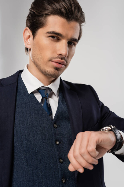 joven hombre de negocios con reloj de pulsera de pie en traje y mirando a la cámara aislada en gris - Foto, imagen