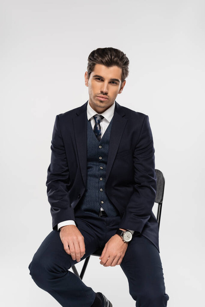 young businessman in formal wear sitting on chair while looking at camera isolated on grey - Foto, imagen