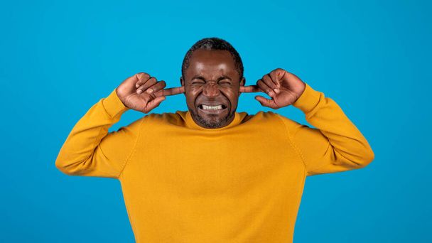 アフリカ系アメリカ人の成人男性は指で耳を差し込む。男性は黄色の深刻な怒りの顔で耳をカバーし、叫んで、歯を食いしばる。ポートレート、スタジオ撮影、怒りの概念、青の背景 - 写真・画像