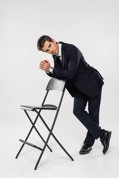 full length of successful businessman in formal wear standing near chair on gray - Foto, Bild