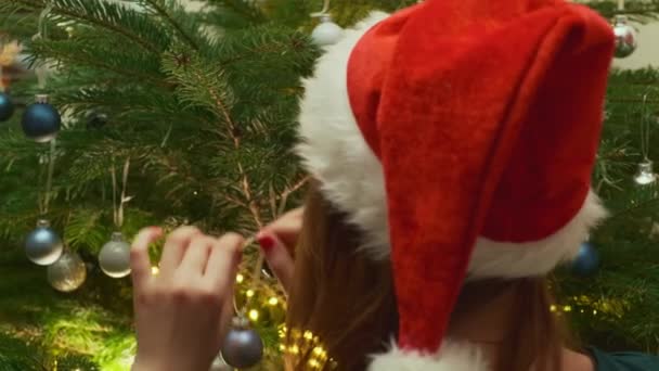 Une fille dans un chapeau de Père Noël accroche une balle bleue sur un arbre de Noël. Décorer le sapin de Noël avec des lumières en arrière-plan.  - Séquence, vidéo