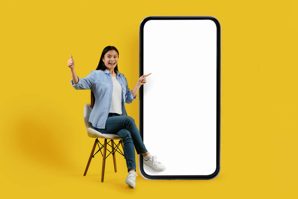 Glad shocked millennial chinese lady student in casual sit on chair, point fingers at big smartphone with empty screen isolated on yellow background. Presentation of new app, huge sale, ad and offer - Photo, Image