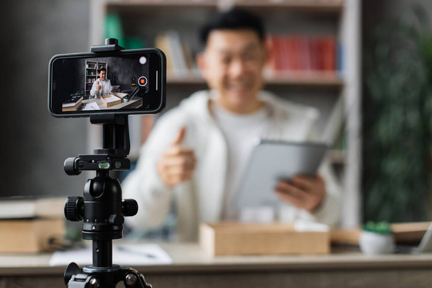 Focus on smart phone screen , asian man recording video on phone camera while unpacking box with new tablet. Young male influencer sharing with subscribers his positive feedback about new order. - 写真・画像