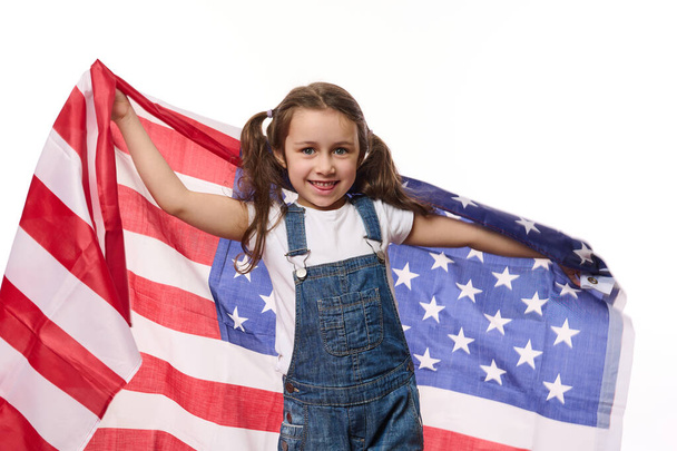 Adorable American child girl in blue denim overalls, carries USA flag, celebrates Independence Day on July 4th. Citizenship, immigration, emigration, winning the green card lottery and freedom concept - Photo, Image