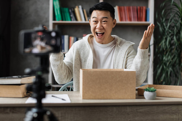 Excited overjoyed young asian male blogger unpacking gift boxes presents from companies and doing live streaming. Attractive man using smart phone camera for creating content. - Foto, Imagem