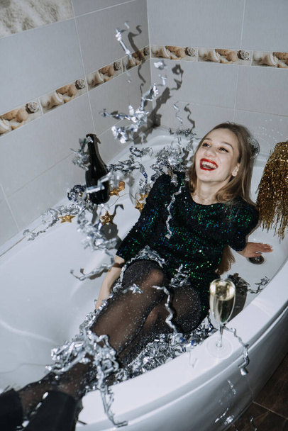 Bathtub Party Day, Party Bathroom Decor. New Years or Birthday Party in Bathtub. Happy girl in little black dress holding champagne and confetti while sitting in bathtub at party. - Zdjęcie, obraz