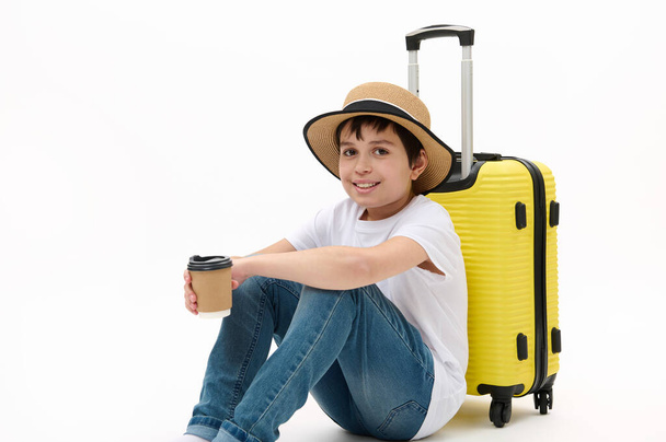Szczęśliwy nastoletni chłopiec podróżnik w luźnym ubraniu i słomkowym kapeluszu, z żółtą walizką i napojem na wynos w jednorazowym eko papierowym kubku, czekający na wejście na pokład samolotu, odizolowany na białym tle. Wolna przestrzeń reklamowa. - Zdjęcie, obraz