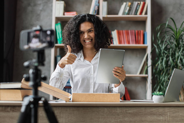 Multicultural woman recording video on phone camera while unpacking box with new wireless tablet. Female influencer sharing with subscribers her positive feedback about new order. - Foto, Bild