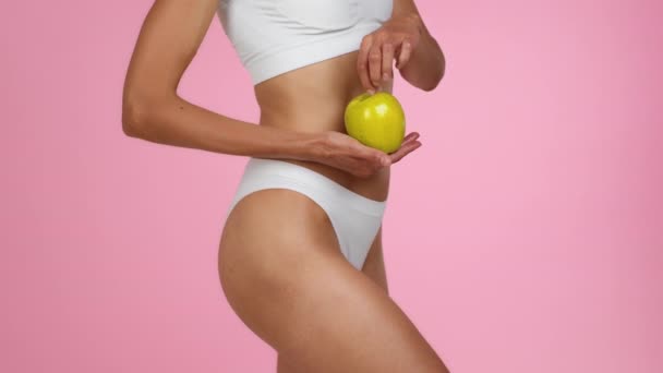 Здоровое спортивное питание. Крупный план неузнаваемой здоровой женщины в нижнем белье, держащей в руках свежее органическое зеленое яблоко, позирующей на розовом фоне студии, замедленной съемки - Кадры, видео