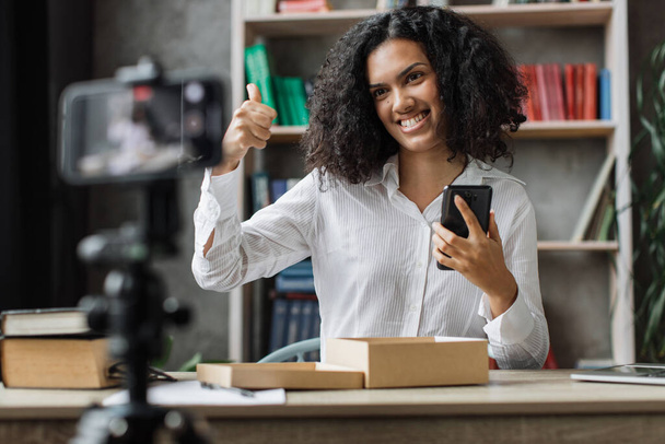 Pleasant multinational woman filming video on modern phone camera while opening parcel box with new smartphone showing thumb up. Concept of people, technology and blogging. - Photo, Image