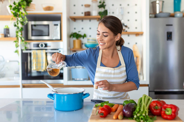 Ευτυχισμένη νεαρή γυναίκα μαγείρεμα γευστικό δείπνο σε ένα δοχείο στέκεται στη σύγχρονη κουζίνα στο σπίτι. Νοικοκυρά Προετοιμασία Υγιεινής Τροφίμων Χαμογελώντας. Οικιακό και Διατροφή. Έννοια συνταγών δίαιτας - Φωτογραφία, εικόνα