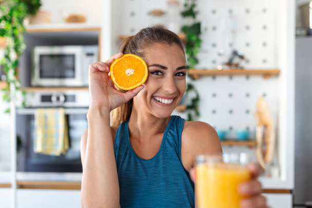 Όμορφη νεαρή γυναίκα πίνει φρέσκο χυμό πορτοκαλιού στην κουζίνα. Υγιεινή διατροφή. Ευτυχισμένη νεαρή γυναίκα με ένα ποτήρι χυμό και πορτοκάλι στο τραπέζι της κουζίνας. - Φωτογραφία, εικόνα