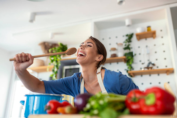 Funny beautiful woman singing into spatula, cooking in modern kitchen, holding spatula as microphone, dancing, listening to music, playful girl having fun with kitchenware, preparing food. - Photo, Image