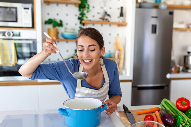 Onnellinen nuori nainen ruoanlaitto makupala pilvi seisoo moderni keittiö kotona. Kotiäiti valmistelee terveellistä ruokaa hymyillen. Kotitalous ja ravitsemus. Ruokavalion reseptit käsite - Valokuva, kuva