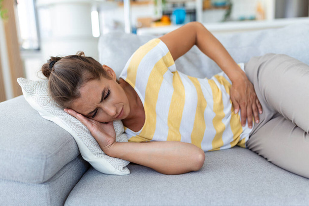 Woman lying on sofa looking sick in the living room. Beautiful young woman lying on bed and holding hands on her stomach. Woman having painful stomachache on bed, Menstrual period - Photo, Image