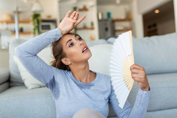 Woman puts head on sofa cushions feels sluggish due unbearable heat, waves hand fan cool herself, hot summer flat without air-conditioner climate control system concept - Foto, Bild