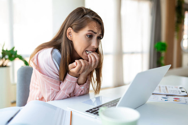 Egy fiatal nő rágja a körmét, miközben otthon laptopozik. Az irodában dolgozó ideges nő ujjait és körmeit harapdálja.. - Fotó, kép