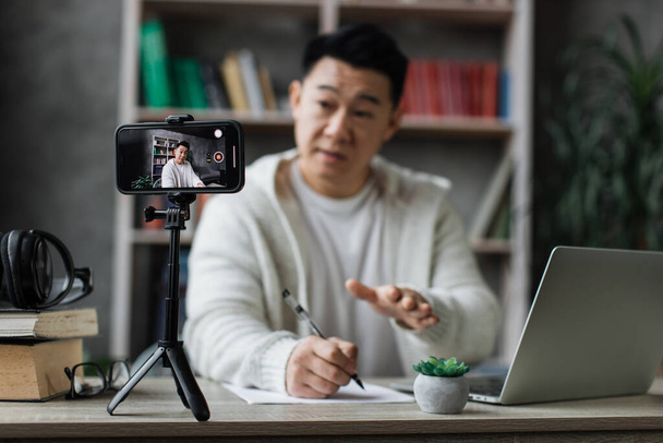 Focus on screen, smiling asian man talking and gesturing in front of modern smartphone camera on tripod, writing report while sitting on background of modern home. - Foto, Bild