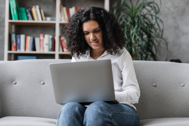 Attractive african american woman in casual clothes sitting on couch and using wireless laptop. Young female browsing internet during leisure time at cozy home. - Foto, afbeelding