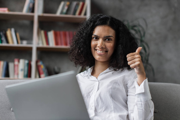 Close up of happy multinational woman in casual clothes sitting on couch and using wireless laptop showing thumb up. Young female browsing internet during leisure time at cozy home. - Foto, Bild