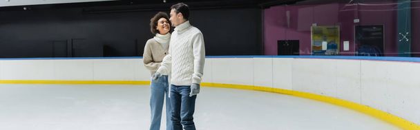 Smiling african american woman in sweater looking at boyfriend on ice rink, banner  - Photo, Image