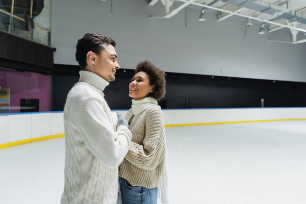 Smiling african american woman looking at boyfriend in sweater on ice rink  - Φωτογραφία, εικόνα