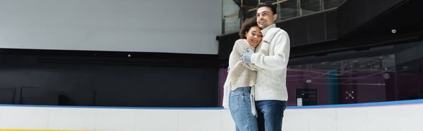 Man in knitted sweater hugging african american girlfriend on ice rink, banner  - Fotó, kép