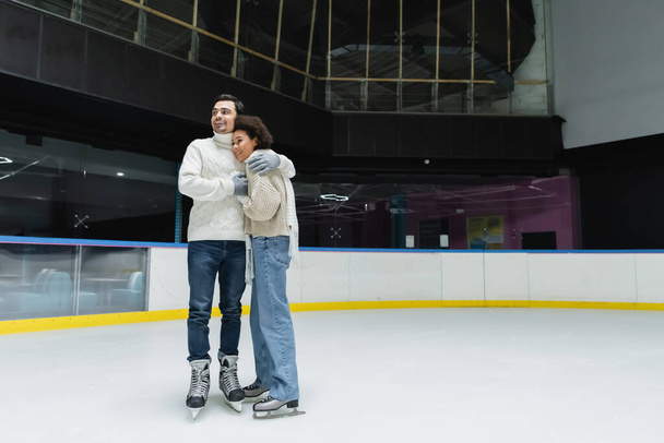 Smiling man hugging african american girlfriend in sweater and scarf on ice rink  - Photo, Image
